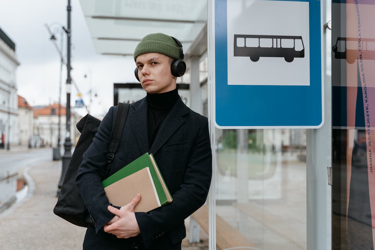 A young man holding books, listening to music, waits at a bus stop in an urban setting.