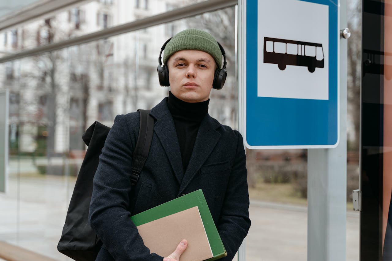 A man in a beanie and headphones holds books while waiting at a bus stop. Urban commuter vibe.